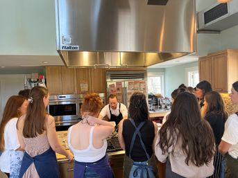 Chef demonstrating a recipe to a group of participants in aprons gathered around a stovetop under a large stainless-steel range hood in a bright kitchen.