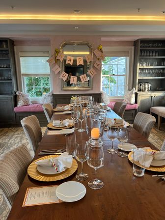 Intimate private dining room set for a birthday dinner — long wooden table with gold chargers, folded napkins, wine glasses and candle centerpiece, pink 'Happy Birthday' banner over a mirror, cushioned window seats and built-in cabinets.