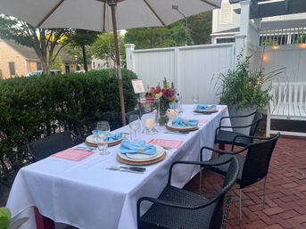 Al fresco backyard patio table set for six on a brick terrace with white tablecloth, blue napkins, glassware, floral centerpiece, umbrella and black wicker chairs.