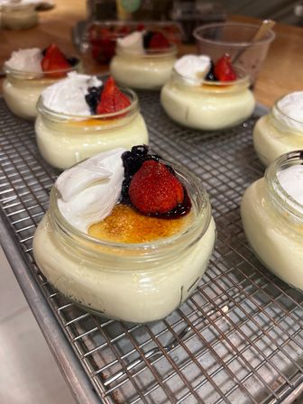 Mini creme brulees in small glass jars with caramelized sugar tops, dollops of whipped cream and fresh strawberries with berry compote, lined up on a metal cooling rack in a kitchen.