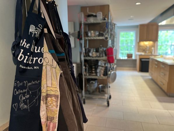 Colorful aprons hanging by a doorway with a stainless-steel rack of pots and pans and a bright modern kitchen with wood cabinets and tile floor