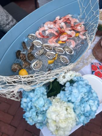 Boat-shaped blue tub of crushed ice holding shucked oysters and cooked shrimp with lemon wedges, draped in a fishing net beside blue and white hydrangea bouquet.