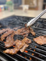 Sizzling marinated beef slices with charred grill marks on a hot grill, metal tongs lifting a juicy seared piece above glowing coals and smoke.