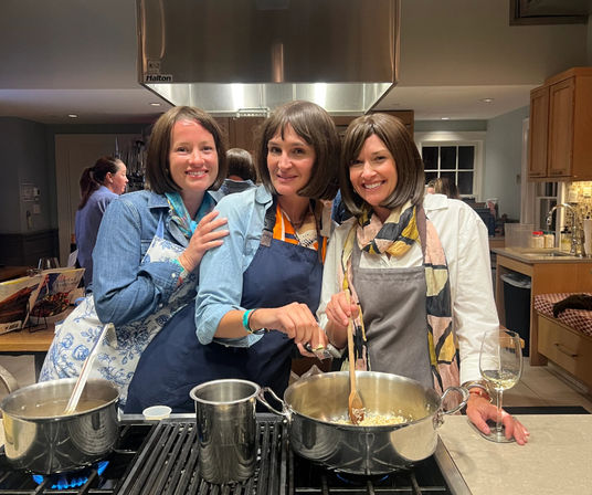 Three friends in aprons cheerfully cooking in a modern home kitchen — stirring a large pot on the stovetop with pans simmering and a glass of white wine on the counter.