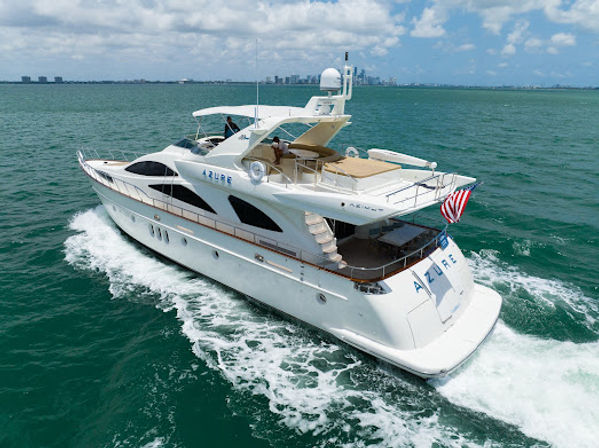 Aerial view of a luxury white motor yacht cruising turquoise waters, leaving a foamy wake, with sun deck seating and an American flag, coastal city skyline on the horizon under a partly cloudy sky.