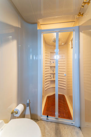 Compact cream-colored marine-style bathroom with sliding glass shower doors, curved built-in shelves, chrome fixtures and a wooden slatted floor mat, alongside a toilet with a gold-tone toilet paper holder.
