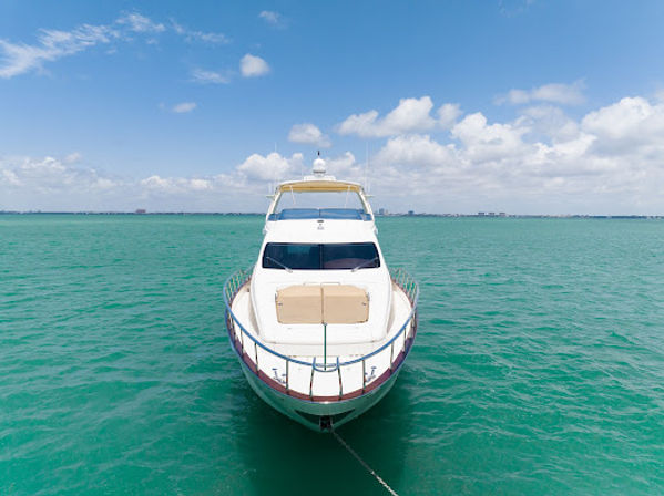 Aerial view of a luxury white yacht moored in turquoise tropical sea, bow facing the camera with anchor line visible under a bright blue sky with fluffy clouds.