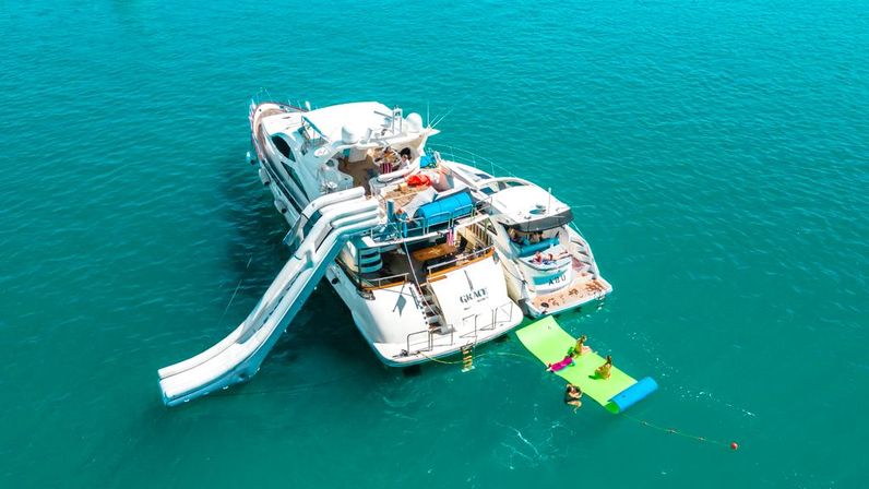 Luxury white motor yacht anchored in clear turquoise sea with triple water slides, an inflatable green swim mat, and people lounging and swimming off the stern.