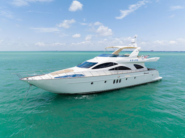 Aerial view of a gleaming white luxury yacht anchored in turquoise tropical waters under a bright blue sky with scattered clouds