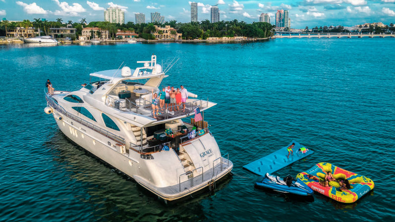 Luxury white yacht anchored in turquoise tropical water by a palm-lined waterfront and city skyline; people on deck with a jet ski, floating mat and colorful inflatable tube on a sunny day