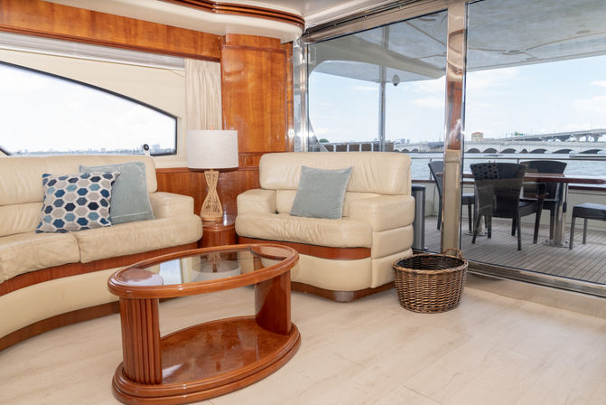 Bright luxury yacht salon with cream leather sofas, wooden trim, glass coffee table and lamp, sliding glass doors to an aft deck dining area and waterfront view of a bridge.
