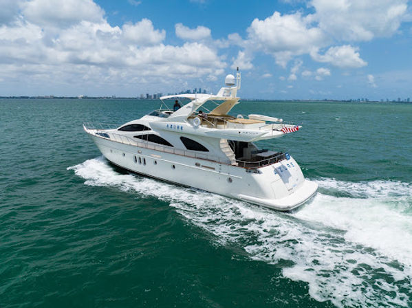 White luxury motor yacht cruising through turquoise ocean, leaving a foamy wake with a distant city skyline under a bright blue, partly cloudy sky.