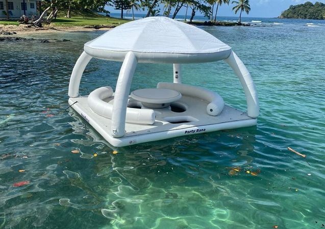 White inflatable floating lounge with canopy, circular bench and small table drifting on clear turquoise tropical water near a palm-lined beach