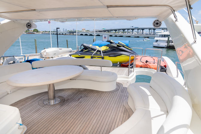 Sunlit yacht aft deck with curved white seating and round table, two yellow-black jet skis on the swim platform, orange life raft, marina waters and a bridge in the background
