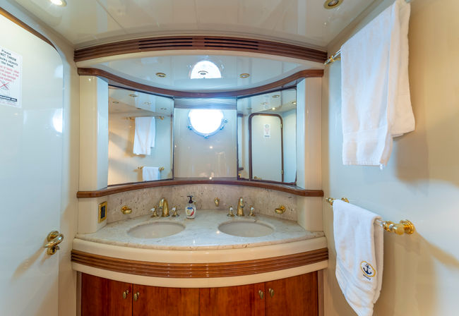 Onboard luxury yacht bathroom (head) with twin marble sinks, polished gold fixtures, curved wood trim, mirrored vanity, round porthole window and white towels on gold rails.