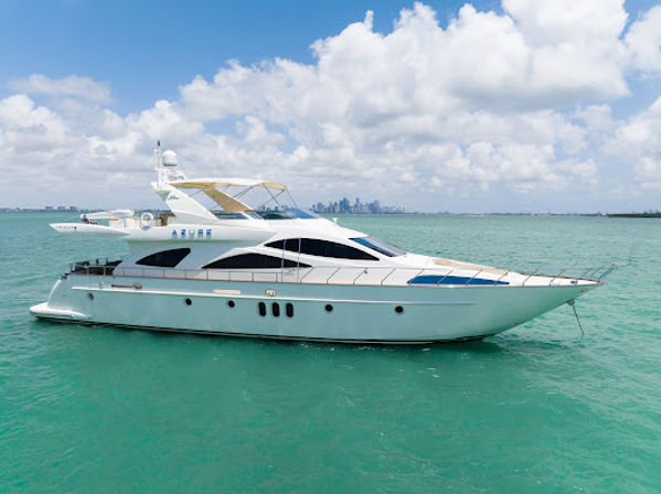 Sleek white luxury yacht in turquoise water off a coastal city skyline under blue sky and fluffy clouds