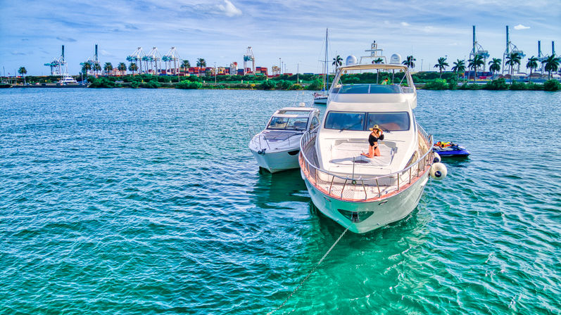 Luxury yacht anchored in turquoise tropical harbor with a woman sunbathing on the bow, a smaller boat and jet ski nearby, palm trees and port cranes on the shoreline