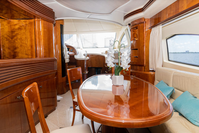 Luxury yacht interior dining area with glossy mahogany table and chairs, white orchid centerpiece, cream leather sofa with teal pillows and large windows showing water and distant city skyline.