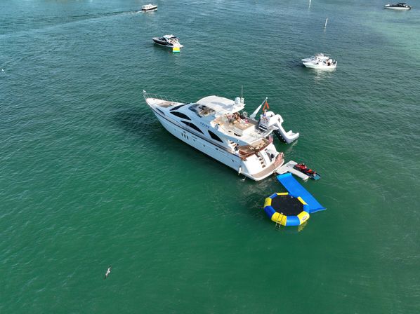 Aerial view of a luxury white yacht anchored in green-blue coastal water with a waterslide, blue ramp, inflatable yellow-and-blue trampoline and a red jet ski nearby, surrounded by smaller boats.