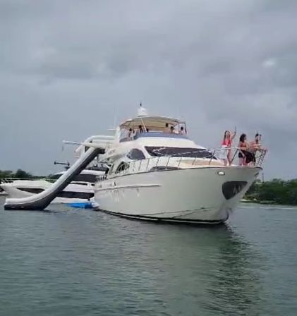 Luxury white yacht anchored in calm coastal water under an overcast sky, inflatable water slide and floating platform at the stern, people in swimwear enjoying a yacht party on the bow.