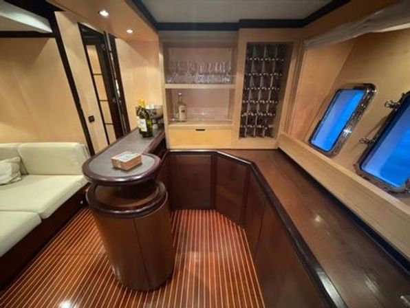 Interior of a luxury yacht salon featuring a curved mahogany bar with wine rack and glassware, teak-and-holly striped flooring, cozy cream sofa, and twin porthole windows showing blue sea.