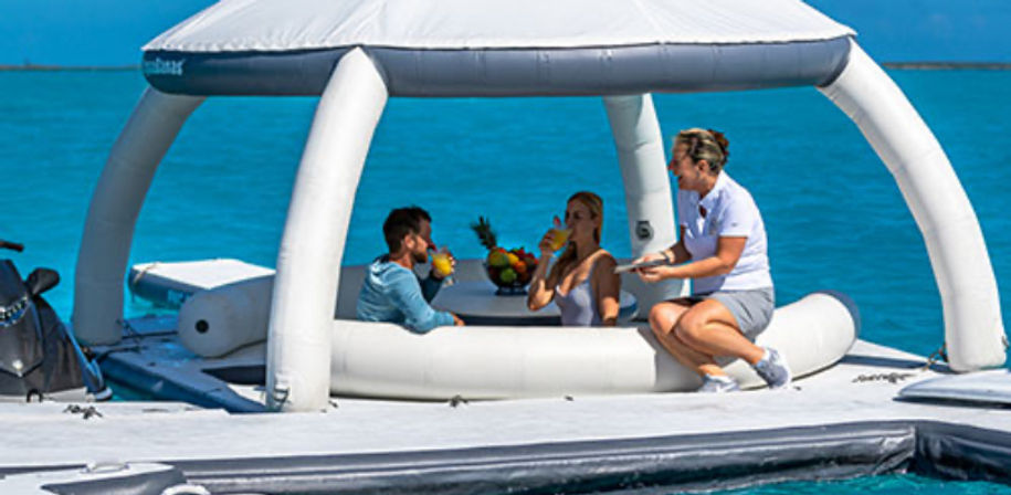 Three people relaxing on a white inflatable floating lounge with canopy in turquoise ocean, two guests sipping tropical drinks at a small table with a fruit bowl while a server hands a drink