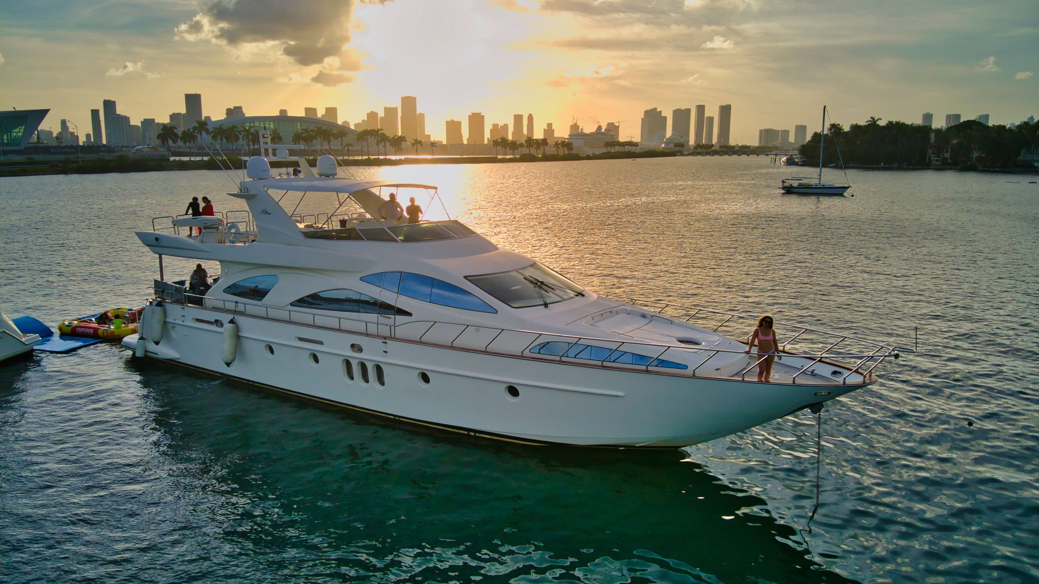 Luxury white yacht anchored in a tropical bay at sunset, city skyline in the background, a person on the bow and inflatable water toys tied alongside.