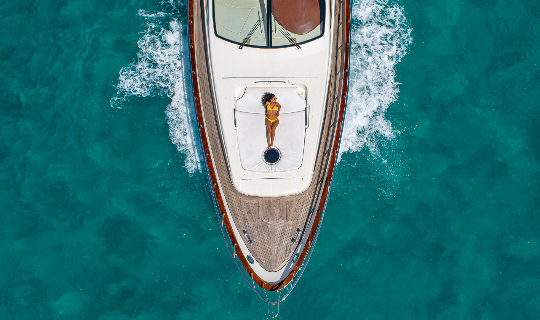 Aerial drone view of a luxury yacht cutting through clear turquoise water with a person in a yellow swimsuit sunbathing on the bow.