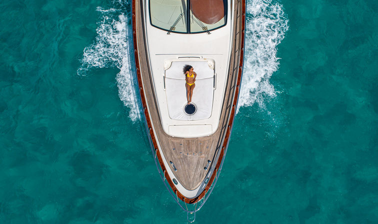 Aerial drone view of a luxury yacht cutting through clear turquoise water with a person in a yellow swimsuit sunbathing on the bow.