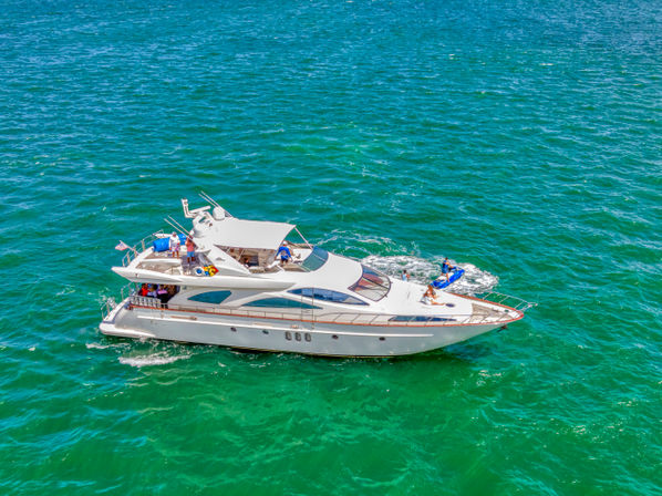 White luxury yacht cruising turquoise ocean on a sunny day, passengers relaxing on deck with a blue jet ski trailing near the bow