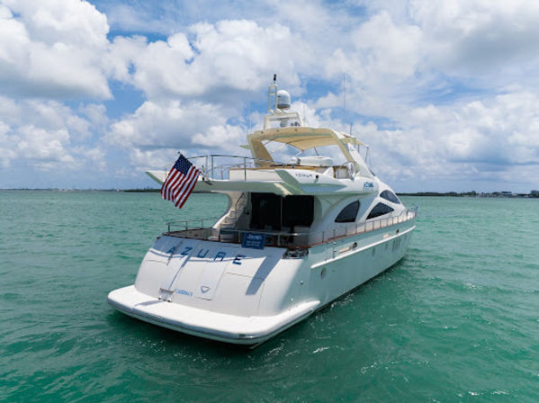 White luxury motor yacht floating on turquoise coastal waters with an American flag at the stern under a sunny, partly cloudy sky and low shoreline on the horizon.