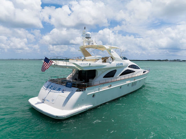 Sleek white luxury motor yacht flying an American flag, floating in turquoise tropical waters off a sunny coastline under a bright blue sky with puffy clouds.