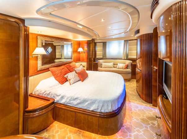 Luxurious yacht master stateroom with curved wood cabinetry, rounded bed dressed in a white quilt and orange accent pillows, small sofa, built-in TV, ornate mirror and warm recessed lighting.