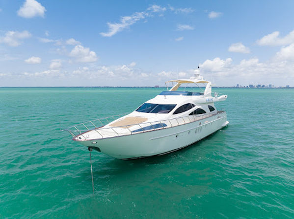 Drone view of a sleek white luxury yacht anchored in clear turquoise sea under a sunny blue sky with fluffy clouds and a distant coastal skyline.