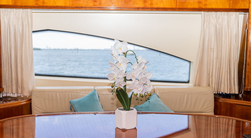 White orchid centerpiece on a glossy wood dining table inside a yacht, cream sofa with teal pillows and a calm ocean view through a large window