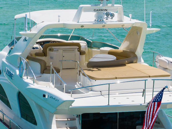 Luxury white motor yacht aft sun deck with tan cushioned loungers and circular seating at the helm, floating on turquoise coastal water with an American flag at the stern