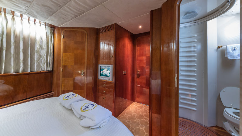 Luxurious yacht cabin with glossy teak wood paneling, curtained window, queen bed topped with folded towels showing an anchor motif, built-in TV and storage, and a curved ensuite bathroom with shower and marine toilet.