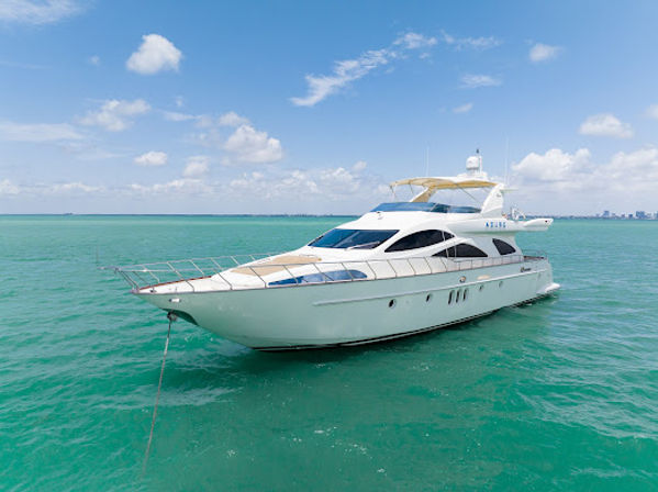 Sleek luxury white motor yacht anchored in clear turquoise tropical coastal waters under a sunny blue sky with a distant shoreline on the horizon