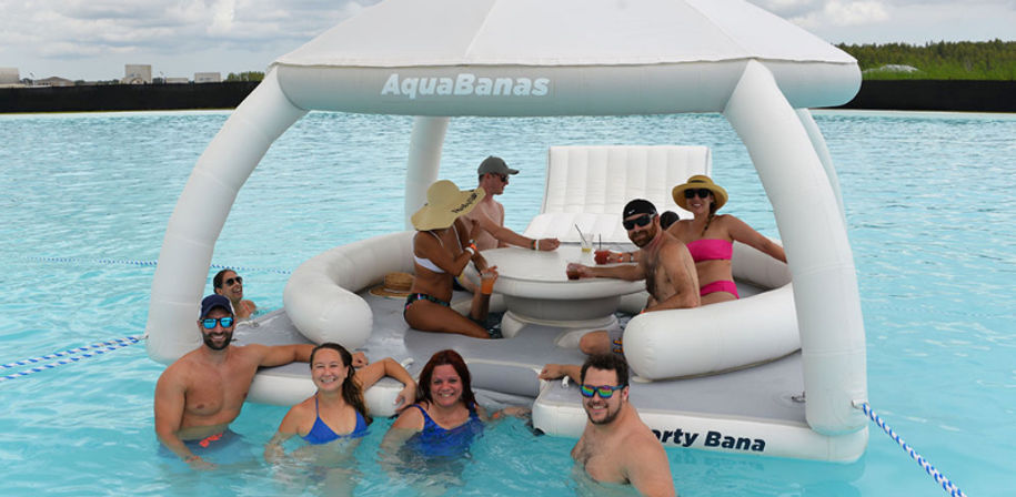 Group of friends in swimsuits smiling and sipping drinks on a white inflatable floating lounge with canopy in a large outdoor turquoise pool