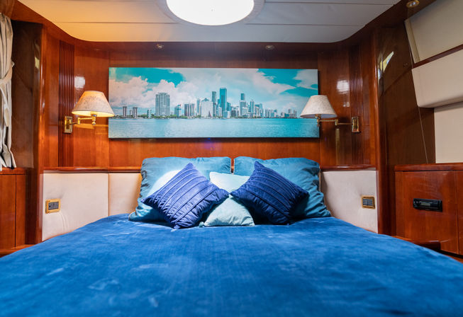 Blue-themed luxury yacht cabin stateroom with plush pillows, polished wood paneling, bedside lamps, and city skyline artwork above the bed.