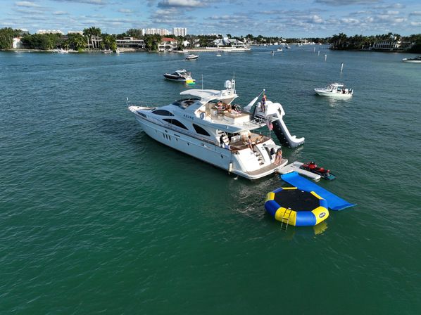White luxury yacht anchored in a Florida coastal inlet with palm-lined waterfront homes, an inflatable slide, blue-and-yellow water trampoline, jet skis, and nearby small boats under a sunny, partly cloudy sky.