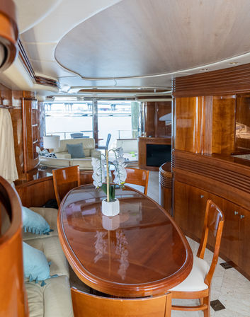 Sunlit luxury yacht interior dining area with glossy oval wooden table, white orchid centerpiece, cream leather seating and marina view through large windows.