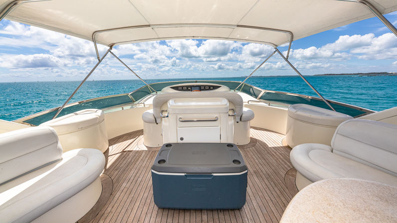 Luxury yacht deck with teak flooring and white curved seating, a blue cooler in the center under a canvas bimini, turquoise ocean and partly cloudy blue sky on the horizon.