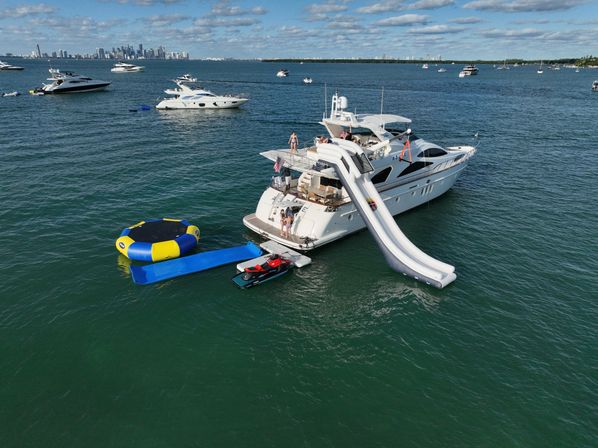 Luxury white yacht anchored with a giant inflatable water slide, blue-and-yellow water trampoline and jet ski on calm coastal waters, other boats nearby and a distant city skyline under a partly cloudy sky.