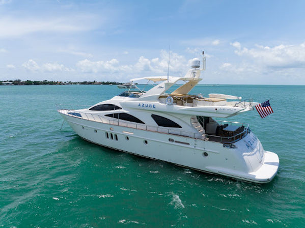Aerial view of a white luxury motor yacht cruising turquoise coastal waters under a sunny blue sky, American flag flying from the stern.
