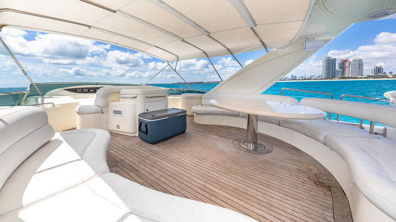 Luxury yacht deck with curved white seating, teak flooring, shaded canopy and round table, portable cooler, overlooking turquoise ocean and a distant coastal city skyline under a bright blue sky