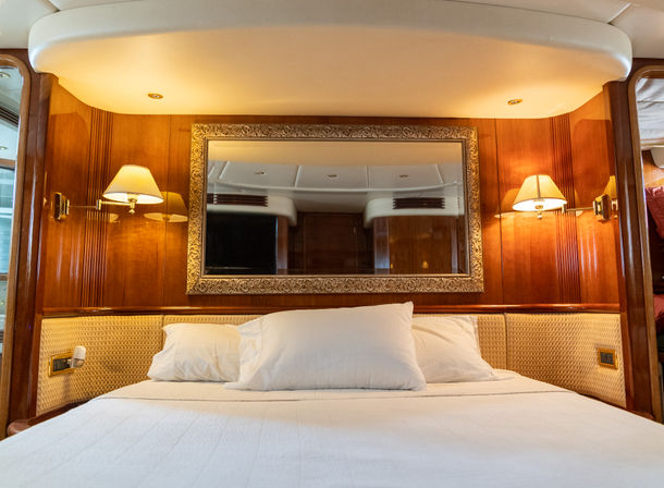 Luxury yacht stateroom with a large bed dressed in white linens and pillows, warm wood-paneled walls, an ornate mirror above the headboard, and wall sconces casting soft light — cozy nautical cabin interior.