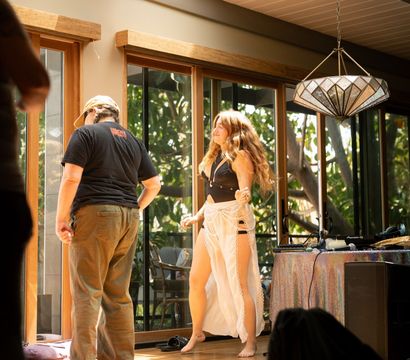 Sunlit room with skylights and wooden sliding glass doors; barefoot woman in a boho sheer skirt and black top dances by a DJ table while a casually dressed person stands on orange yoga mats, laid-back indoor garden vibe.