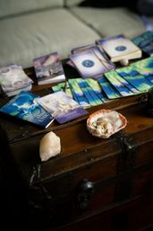 Colorful oracle cards, guidebooks and a seashell holding healing crystals arranged on a vintage wooden chest — cozy spiritual reading setup.