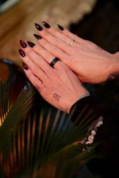 Pair of hands pressed together showing dark red-to-black stiletto nails with tiny gems, a black ring and a small "555" hand tattoo, framed by palm leaves in warm moody lighting.